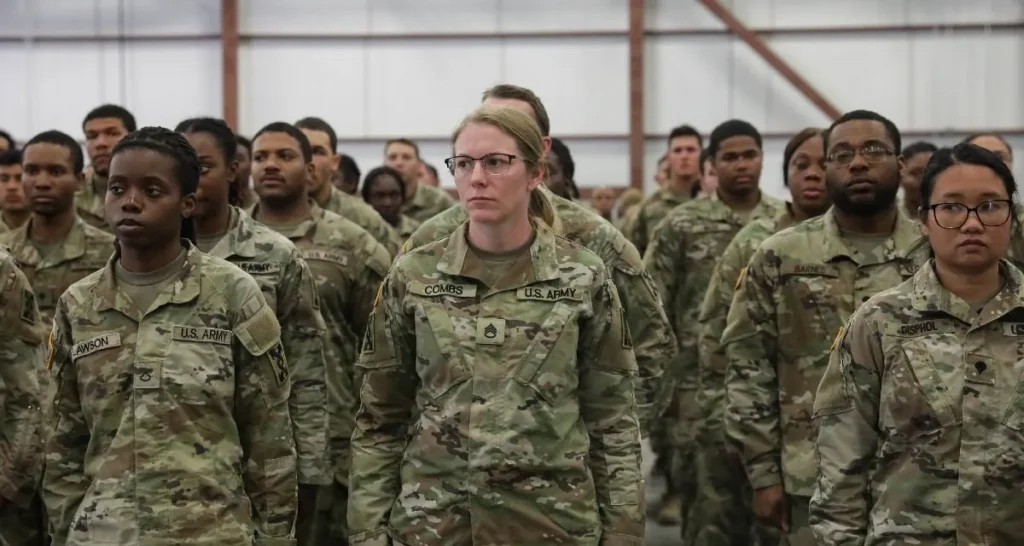 Group of U.S. Army soldiers in camouflage uniforms standing at attention in a hangar, facing forward.