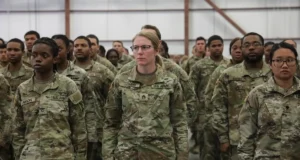 Group of U.S. Army soldiers in camouflage uniforms standing at attention in a hangar, facing forward.