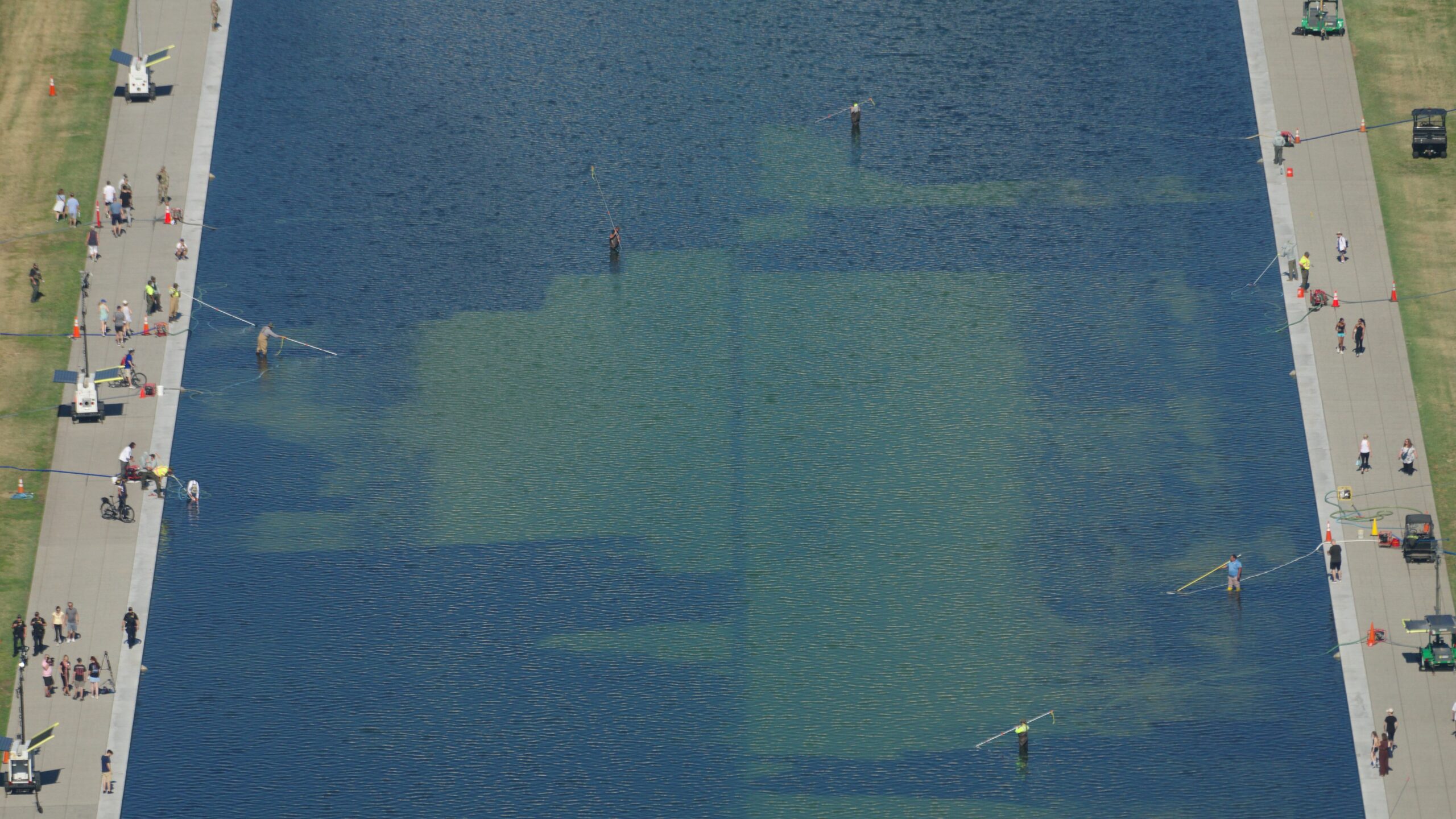 Aerial view of the Lincoln Memorial Reflecting Pool with green water, edges lined by walkways and people strolling, some fishing from the edge.