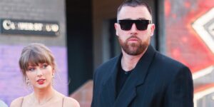 Bearded man in a black suit and sunglasses stands beside a blonde woman with dangling earrings on a city sidewalk.